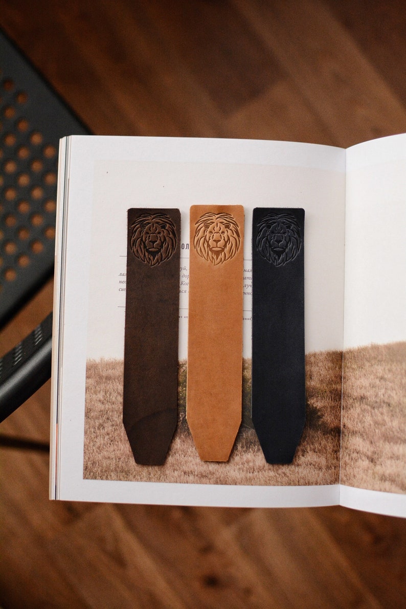 May include: Three rectangular leather bookmarks in dark brown, tan, and black. Each bookmark features a debossed lion head design at the top and a pointed bottom edge. The bookmarks are arranged vertically side by side on an open book page.