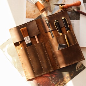 May include: A brown leather roll-up organizer featuring multiple stitched pockets containing two paint brushes, two black pens, a wooden pencil, and a small rectangular eraser. The organizer is laid flat, showcasing its smooth texture and durable construction.