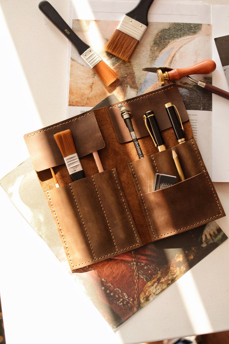 May include: A brown leather roll-up organizer featuring multiple stitched pockets containing two paint brushes, two black pens, a wooden pencil, and a small rectangular eraser. The organizer is laid flat, showcasing its smooth texture and durable construction.