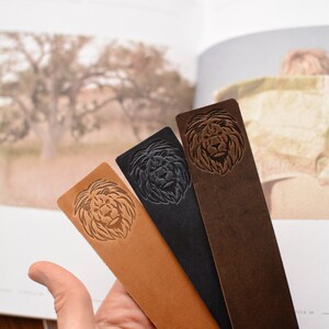 May include: Three rectangular leather bookmarks in tan, black, and dark brown. Each bookmark features a debossed lion head design on the upper portion. The bookmarks are arranged in a fan shape, showcasing the distinct colors and textures.