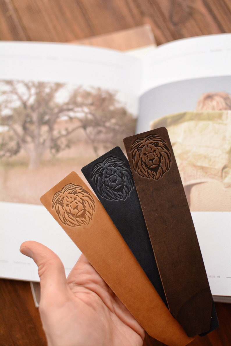 May include: Three rectangular leather bookmarks in tan, black, and dark brown. Each bookmark features a debossed lion head design on the upper portion. The bookmarks are arranged in a fan shape, showcasing the distinct colors and textures.