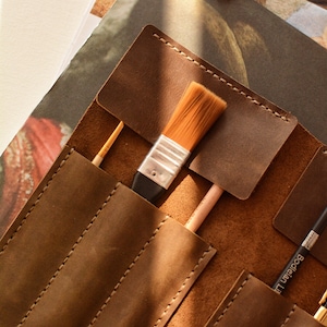 May include: A brown leather roll-up case featuring multiple stitched pockets for organizing art supplies. The case contains a flat-tipped paintbrush with golden bristles, a thin wooden-handled brush, and a black pencil labeled Bodleian Library.