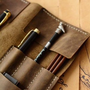 May include: A brown leather pen roll featuring multiple stitched slots for writing instruments. The case holds a black pen with a gold clip, a black pencil with a silver owl topper and the text Bodleian Library, and two brown pencils.