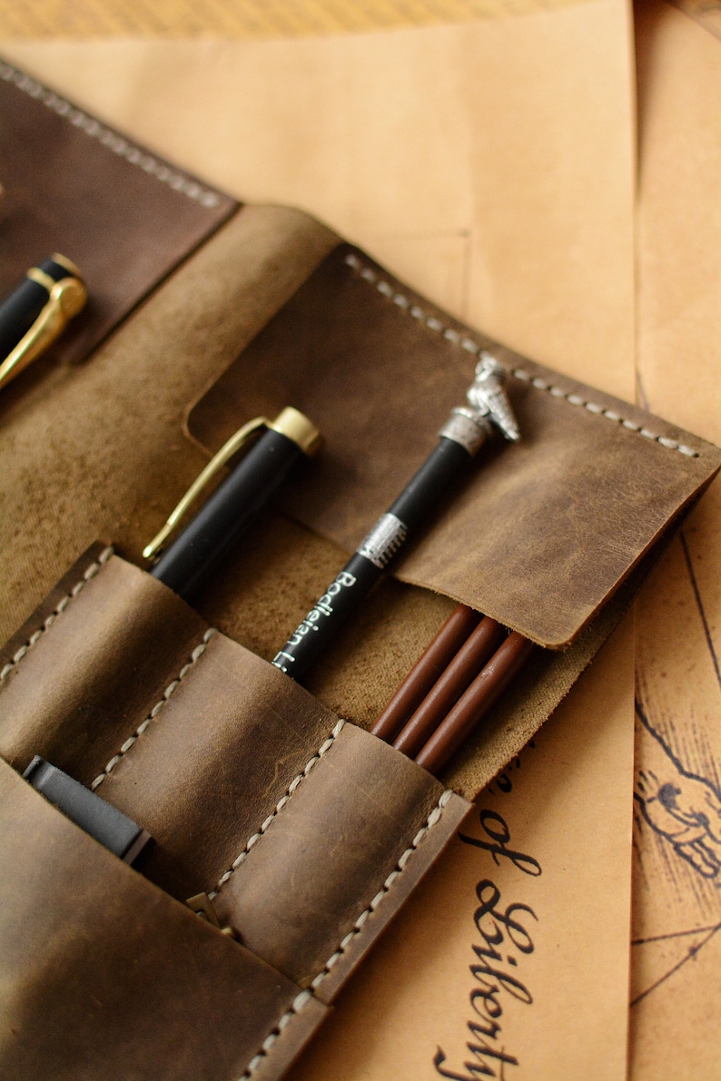 May include: A brown leather pen roll featuring multiple stitched slots for writing instruments. The case holds a black pen with a gold clip, a black pencil with a silver owl topper and the text Bodleian Library, and two brown pencils.