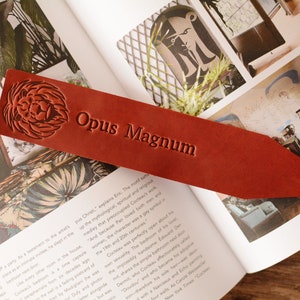 May include: A rectangular, reddish-brown leather bookmark with a pointed end. The surface features an embossed lion head graphic on the left side and the text Opus Magnum embossed in the center, with a smooth, matte finish and clean edges.