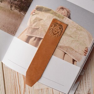 May include: A tan leather bookmark with a pointed end, featuring a debossed lion head design at the top. The bookmark is rectangular in shape with a smooth surface and is placed vertically across the open pages of a book.