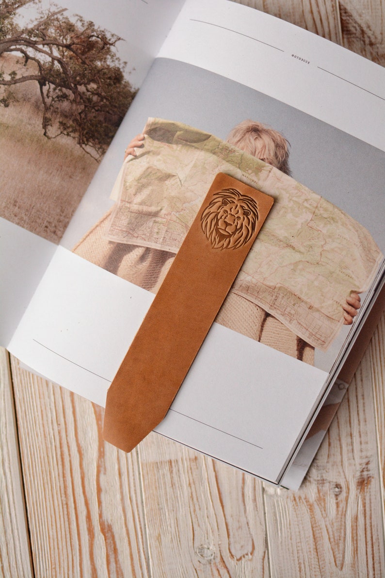 May include: A tan leather bookmark with a pointed end, featuring a debossed lion head design at the top. The bookmark is rectangular in shape with a smooth surface and is placed vertically across the open pages of a book.