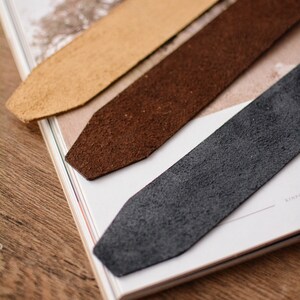 May include: Three rectangular leather bookmarks with pointed ends, arranged diagonally. The bookmarks are colored in light tan, medium brown, and dark charcoal gray, each featuring a textured, suede-like surface finish and a clean, straight-cut edge.