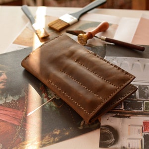 May include: A rectangular brown leather case featuring visible light-colored stitching along the edges and three vertical stitched lines across the front surface. The leather has a smooth, matte texture and a folded construction with a flap closure.