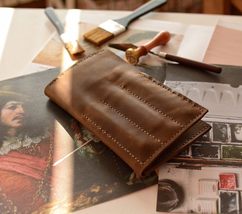 May include: A rectangular brown leather case featuring visible light-colored stitching along the edges and three vertical stitched lines across the front surface. The leather has a smooth, matte texture and a folded construction with a flap closure.
