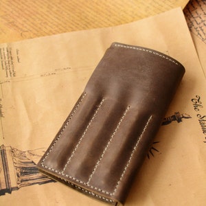 May include: A dark brown leather pen case featuring three vertical stitched slots for individual writing instruments. The case has a smooth, matte finish with visible light-colored stitching along the edges and between each of the three rounded compartments.