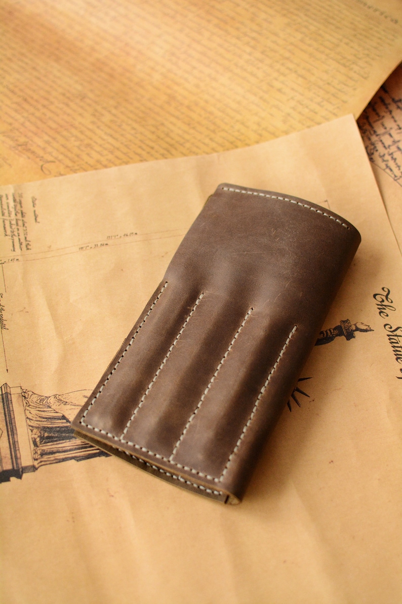 May include: A dark brown leather pen case featuring three vertical stitched slots for individual writing instruments. The case has a smooth, matte finish with visible light-colored stitching along the edges and between each of the three rounded compartments.