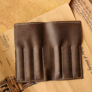 May include: A dark brown leather pen roll featuring four vertical slots for writing instruments. The rectangular case is constructed with visible light-colored stitching along the edges and between each individual pen compartment, creating a structured design for organizing pens.