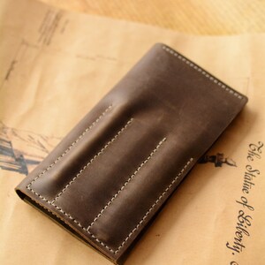 May include: A dark brown leather pen case featuring three vertical stitched slots for individual writing instruments. The rectangular case has visible white stitching along the edges and between each slot, creating a structured design for holding pens.