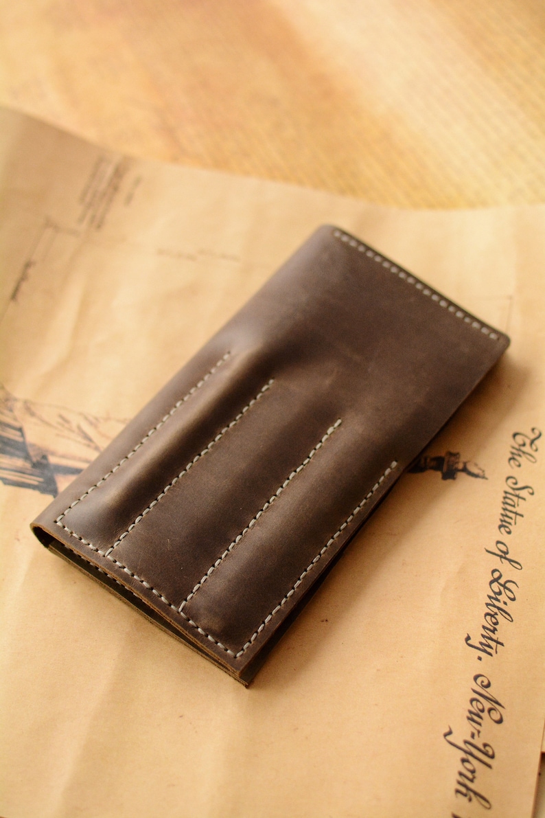 May include: A dark brown leather pen case featuring three vertical stitched slots for individual writing instruments. The rectangular case has visible white stitching along the edges and between each slot, creating a structured design for holding pens.