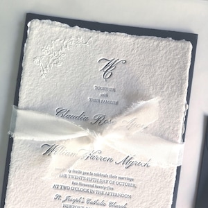 May include: Elegant wedding invitation with a textured white paper and a navy blue border. The invitation includes the names Claudia Rose Apor and William Warren Myrinh, along with event details. A white ribbon is tied around the invitation.