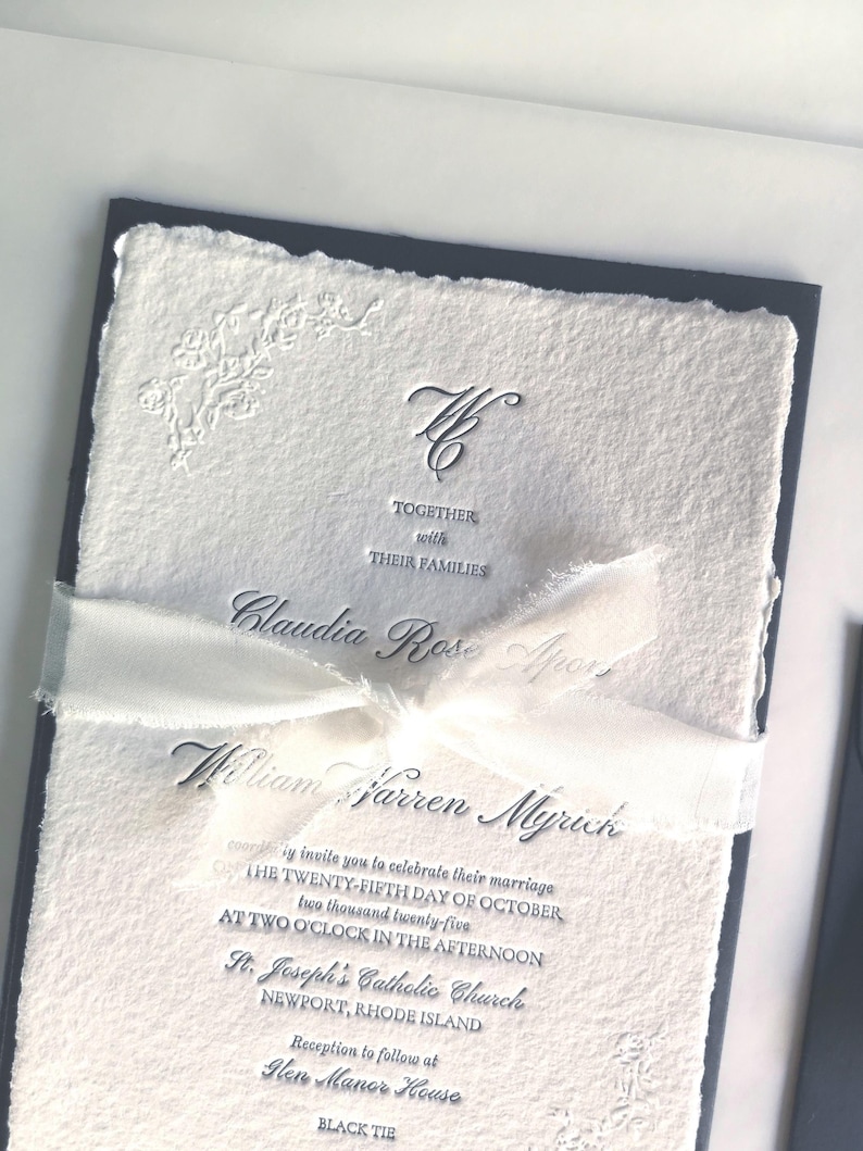 May include: Elegant wedding invitation with a textured white paper and a navy blue border. The invitation includes the names Claudia Rose Apor and William Warren Myrinh, along with event details. A white ribbon is tied around the invitation.