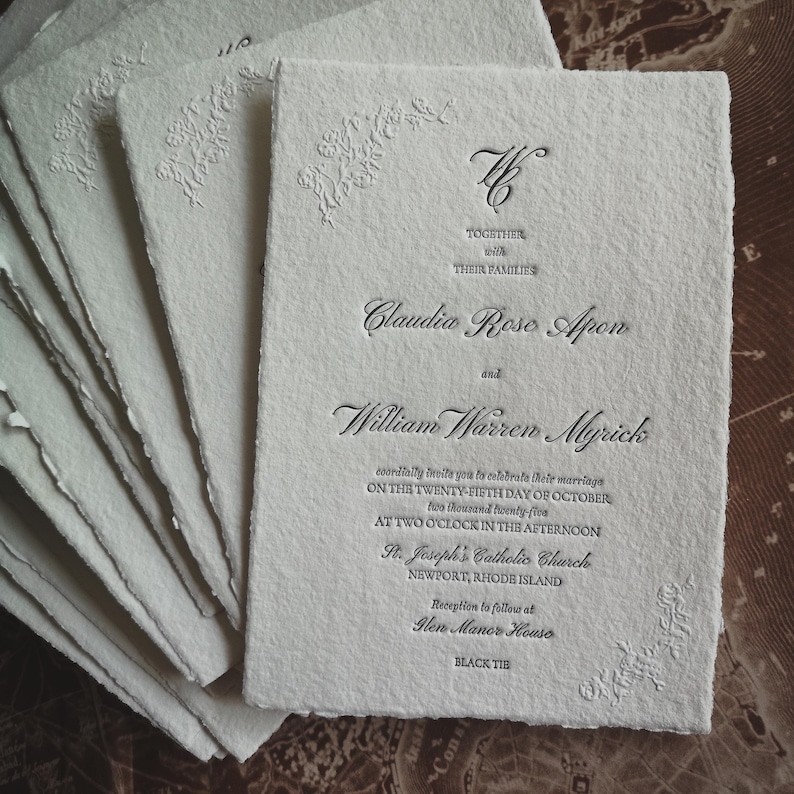 May include: A stack of off-white wedding invitations with deckled edges. The invitations feature embossed floral corner designs and elegant script with the names Claudia Rose Apon and William Warren Myrick. The text includes the wedding date and location.