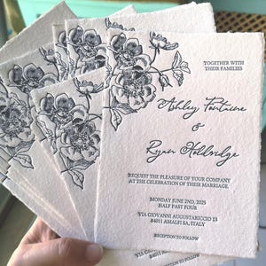 Könnte beinhalten: Eine Reihe cremefarbener Hochzeitseinladungen mit einem floralen Design in schwarzer Tinte. Die Einladungen enthalten die Namen Ashley Fantrine und Ryan Holdridge sowie Datum und Ort der Hochzeit in Amalfi, Italien.