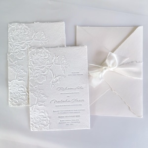 May include: Elegant white wedding invitations with embossed floral designs. The invitation suite includes a card with the names "Raheem Ali" and "Maleiha Nazir", an envelope, and a ribbon. The text reads "Request the pleasure of your company to celebrate their wedding".