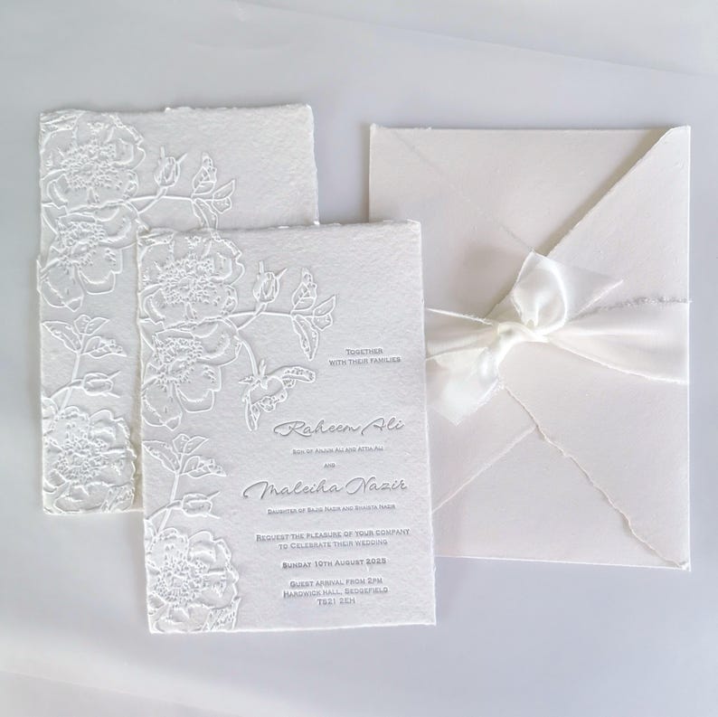 May include: Elegant white wedding invitations with embossed floral designs. The invitation suite includes a card with the names "Raheem Ali" and "Maleiha Nazir", an envelope, and a ribbon. The text reads "Request the pleasure of your company to celebrate their wedding".