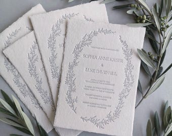 May include: Off-white wedding invitation cards with deckled edges, featuring a debossed silver botanical wreath surrounding text that reads Sophia Anne Kaiser and Luke David Hill, together with our families, joyfully invite you.