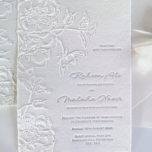 May include: White wedding invitation with embossed floral design. The invitation includes the names Raheem Ali and Maleiha Nazir, along with the wedding date, time, and location. The text "Together with their families" is also included.
