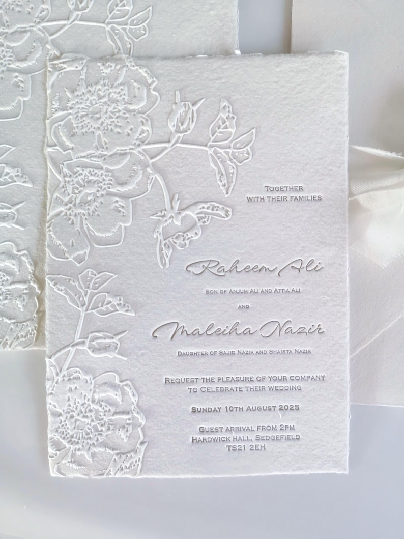 May include: White wedding invitation with embossed floral design. The invitation includes the names Raheem Ali and Maleiha Nazir, along with the wedding date, time, and location. The text "Together with their families" is also included.