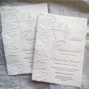 May include: Two off-white wedding invitations with embossed floral designs. The invitations feature the names "Raheem Ali" and "Maleiha Nazir", along with the wedding date and location. The paper has a textured, handmade appearance.