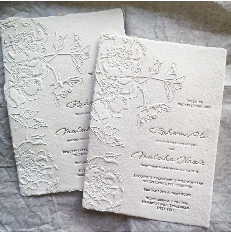 May include: Two off-white wedding invitations with embossed floral designs. The invitations feature the names "Raheem Ali" and "Maleiha Nazir", along with the wedding date and location. The paper has a textured, handmade appearance.