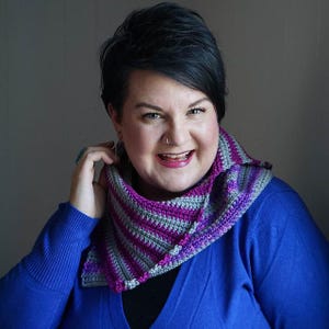 May include: A handmade crochet scarf in shades of purple, gray, and magenta. The scarf features a striped pattern and is worn around the neck. The person in the image is wearing a blue sweater.
