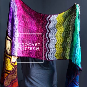 Può includere: Uno scialle all'uncinetto colorato con un motivo a onde, con uno spettro di colori dal viola al blu. Il testo "CROCHET PATTERN" è visualizzato in un quadrato bianco. Lo scialle è tenuto in alto, mostrando il suo design intricato e le sue tonalità vivaci.