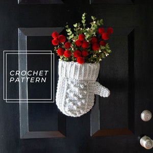 May include: A gray crocheted mitten with a red berry garland hanging on a black door. The text "CROCHET PATTERN" is visible in the image.