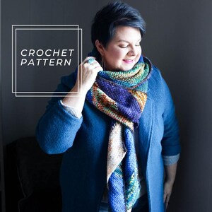 Può includere: Una donna indossa un cappotto blu e una sciarpa all'uncinetto colorata. La sciarpa è a forma triangolare con una varietà di colori, tra cui blu, verde, arancione e bianco. Il testo "CROCHET PATTERN" è scritto in un riquadro bianco sull'immagine.