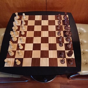 Könnte beinhalten: Ein Schachbrett aus Holz mit schwarzen und braunen Feldern. Die Schachfiguren sind in der Startposition für eine Schachpartie aufgestellt. Das Schachbrett steht auf einem schwarzen Tisch mit abgerundeter Kante. Der Tisch steht zwischen zwei beigen Stühlen mit Knopfpolsterung.