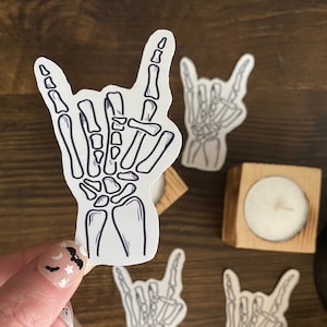 May include: White stickers featuring a skeleton hand in the "rock on" gesture. The stickers have a dark blue outline and are scattered on a wooden surface. The image also includes small wooden candle holders with lit candles.