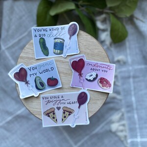 May include: Four colorful stickers with illustrations and text. The stickers feature a pickle and a jar of dill, an avocado and a tomato, two donuts, and two slices of pizza. The text on the stickers reads "You're kind of a big dill", "You grace my world", "I'm donuts about you", and "You stole a pizza my heart".
