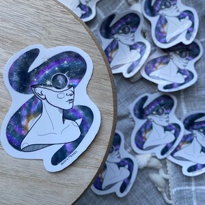 Puede incluir: Un conjunto de pegatinas con una ilustración estilizada de una persona con una galaxia girando alrededor de su cabeza. La persona tiene un tercer ojo en el centro de su frente. Las pegatinas están impresas en papel blanco con un acabado brillante. El texto "Jess's Mess" está escrito en cursiva en la parte inferior de una de las pegatinas.