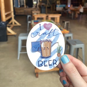 May include: An oval sticker with the words "I craft BEER" and a cartoon image of a beer mug with a smiling face. The sticker has a pink heart and blue accents, with a small potted plant.