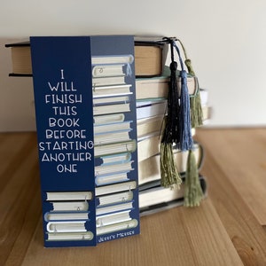 May include: A blue bookmark with the text "I will finish this book before starting another one" and a stack of books illustrated on it. The bookmark is on top of a stack of books with blue, green, and black tassels.