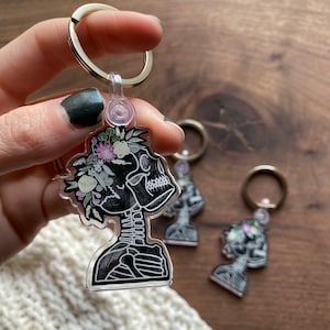 May include: A clear acrylic keychain featuring a black skeleton with a floral crown. The skeleton is depicted in a profile view with its head tilted slightly to the side. The flowers are a mix of white, pink, and green.