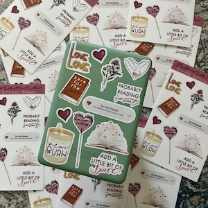 May include: A set of stickers featuring various designs related to reading and romance. The stickers are printed on white paper with a green background. The stickers include illustrations of books, candles, hearts, and text such as "Probably Reading Smut", "Add a Little Bit of Spice", and "Here for the Vibes".
