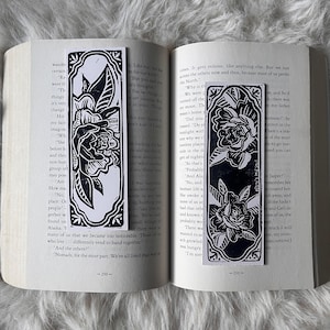 May include: Two black and white floral design bookmarks are displayed on an open book. The bookmarks feature a detailed rose illustration with a decorative border. The book pages are visible in the background.
