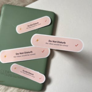 May include: A collection of pink stickers with the text "Do Not Disturb" and "the vibes would be ruined" in a white font. Each sticker has a checkmark and a star graphic. The stickers are on a green electronic device.