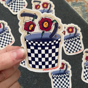 Puede incluir: Una pegatina con un cubo a cuadros blanco y negro con dos flores rojas con caras sonrientes. Las flores sostienen una patineta. La pegatina tiene el texto "Jessi's Messes" en la parte inferior.