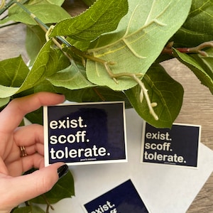 May include: Three blue square stickers with white text that reads "exist. scoff. tolerate." The stickers are on a white surface with green leaves in the background.