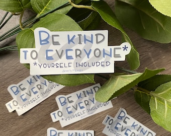 May include: Four white stickers with blue text that reads "Be Kind to Everyone* *Yourself Included". The stickers are on a wooden surface with green leaves in the background.