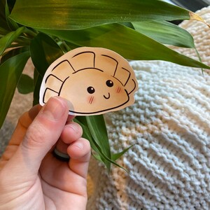 Könnte beinhalten: Ein brauner und weißer Cartoon-Aufkleber mit einer lächelnden Pastete mit einem glücklichen Gesicht. Der Aufkleber befindet sich auf einer weißen gestrickten Oberfläche.