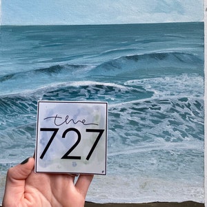 Op de afbeelding: Een witte sticker met een aquarel achtergrond en zwarte tekst die "the 727" luidt. De sticker wordt voor een schilderij van een blauwe oceaan met witte golven gehouden.