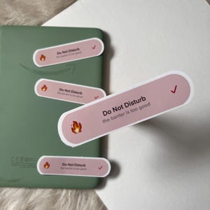 May include: A collection of pink and white stickers with the text "Do Not Disturb" and "the banter is too good". Each sticker features a flame icon and a checkmark. The stickers are shown on a green electronic device.